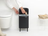 Schwarzer Abfalleimer Bo Hi 7L mit geöffnetem Deckel, von vorne gesehen, neben einer Toilette und einem Tisch mit Handtüchern.