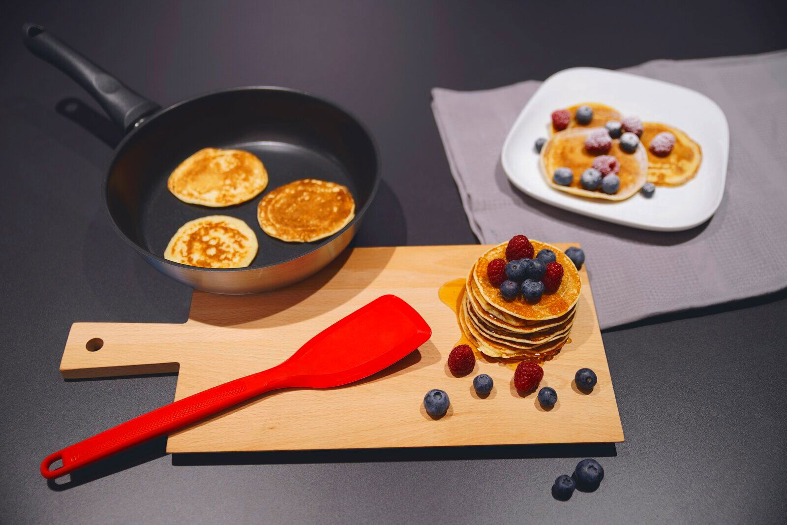Roter Silikon-Wender auf einem Holzbrett, umgeben von Pfannkuchen und Beeren, aus der Vogelperspektive.