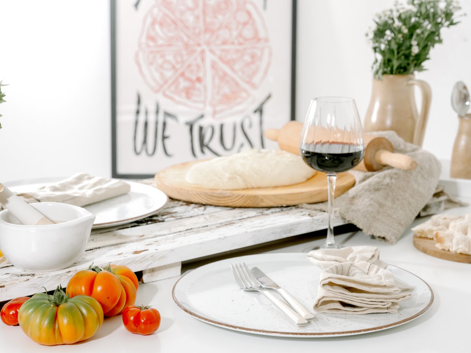 Heller Küchen- und Esstisch mit rustikalem Holzbrett, Teig auf rundem Holzbrett, Nudelholz, Rotweinglas, Teller mit Besteck und Stoffserviette sowie bunten Tomaten im Vordergrund; Perspektive: leicht seitliche Nahaufnahme auf Tischhöhe.
