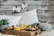 Frühstückstablett aus Holz auf einem Bett mit zwei Gläsern Orangensaft, Croissants, Feigen und Trauben, seitliche Perspektive auf das Arrangement vor rustikaler Holzwand.