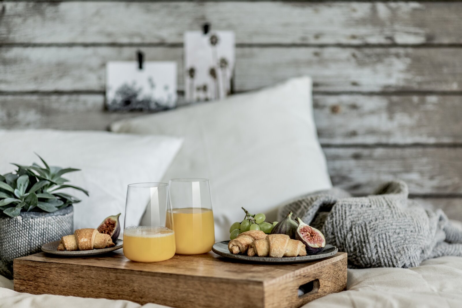 Frühstückstablett aus Holz auf einem Bett mit zwei Gläsern Orangensaft, Croissants, Feigen und Trauben, seitliche Perspektive auf das Arrangement vor rustikaler Holzwand.