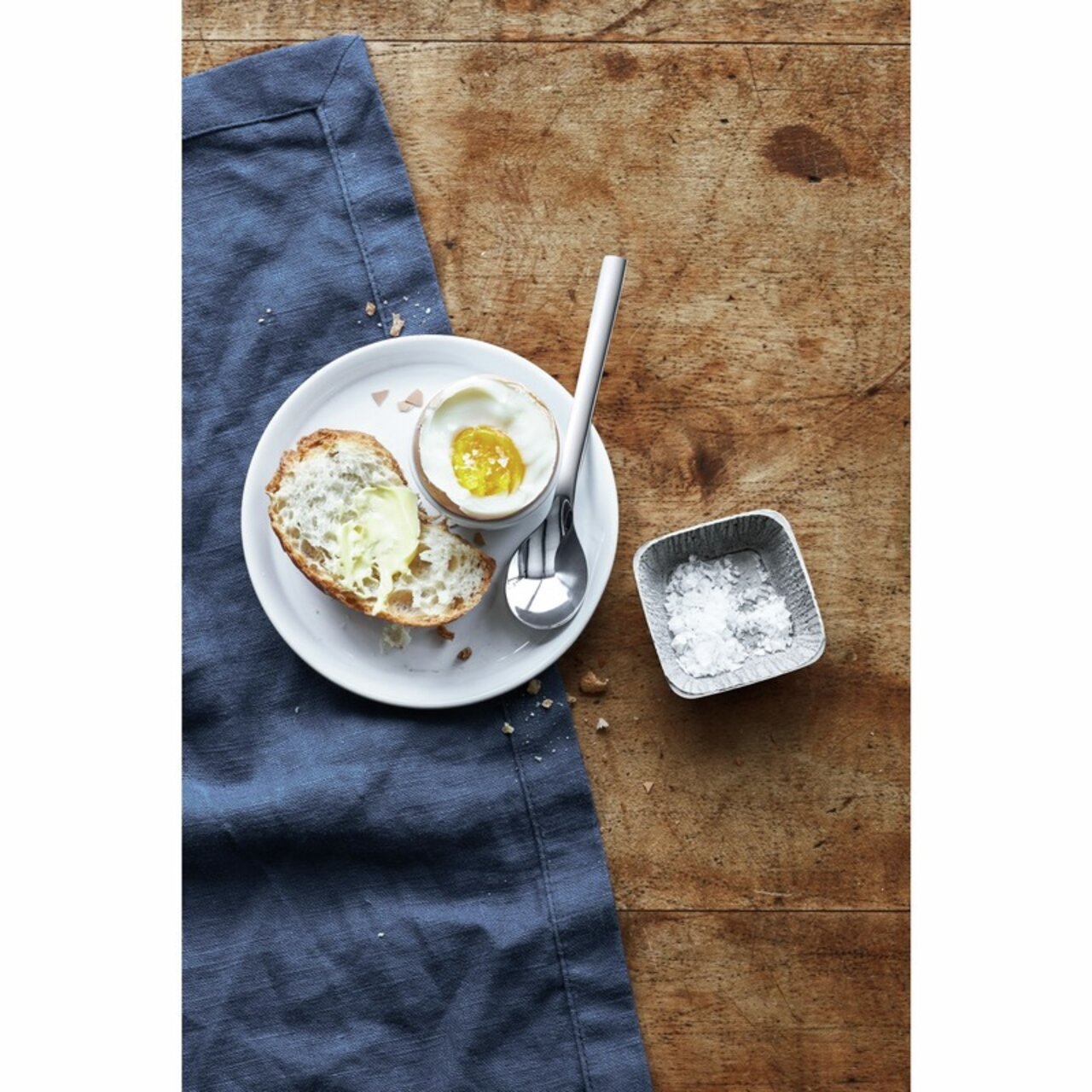 Eierlöffel aus Edelstahl neben einem weichgekochten Ei in einem Eierbecher, auf einem Holztisch von oben fotografiert.