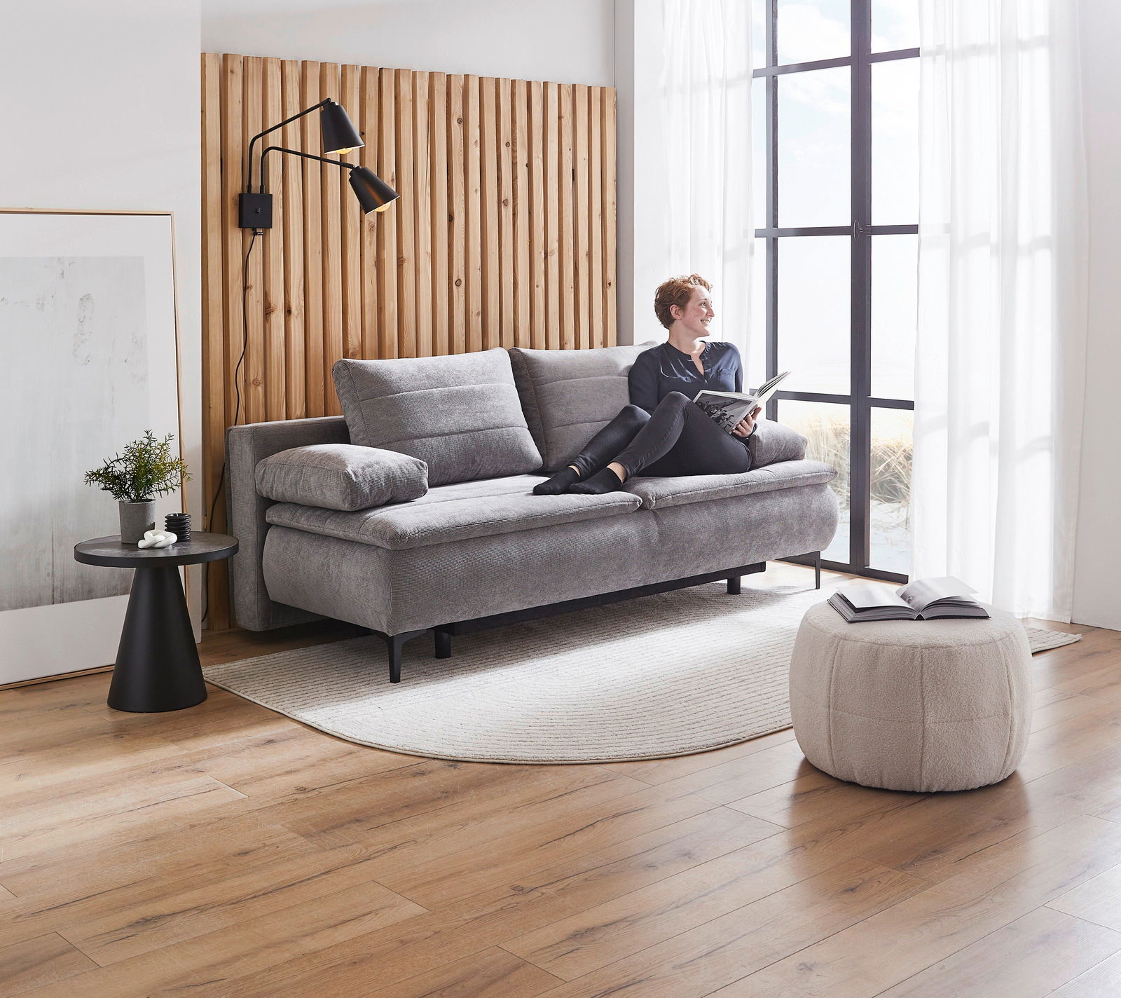 Graues Boxspring-Funktionssofa in modernem Wohnzimmer, seitliche Perspektive, mit einer Person, die darauf sitzt und ein Buch liest.