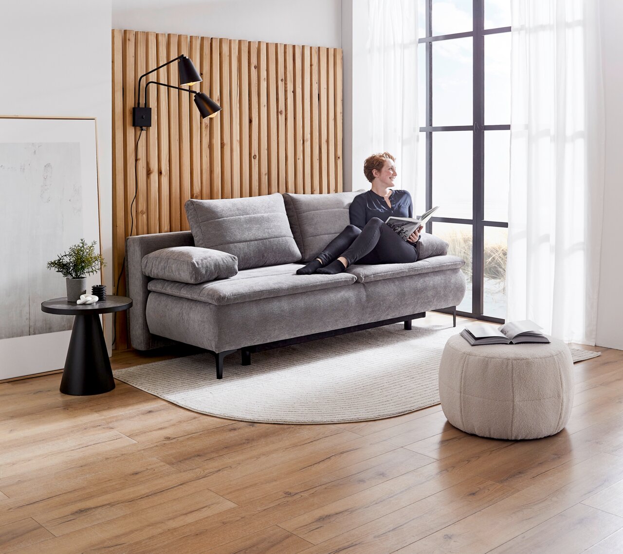 Grijze boxspring-functiebank in een moderne woonkamer, gezien vanaf de zijkant, met iemand die erop zit en een boek leest.