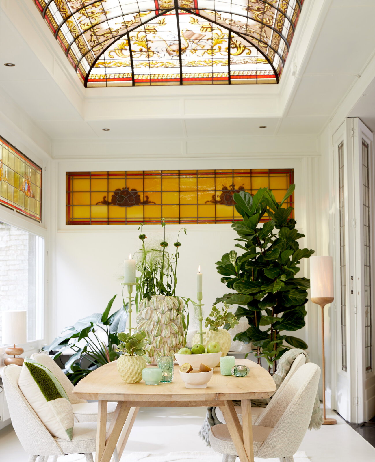 Heller Essbereich mit hellem Holztisch und gepolsterten Stühlen, dekoriert mit grünen Vasen, Kerzen und Obst, im Hintergrund große Zimmerpflanze und Buntglasfenster; Perspektive: frontale Aufnahme auf Augenhöhe.
