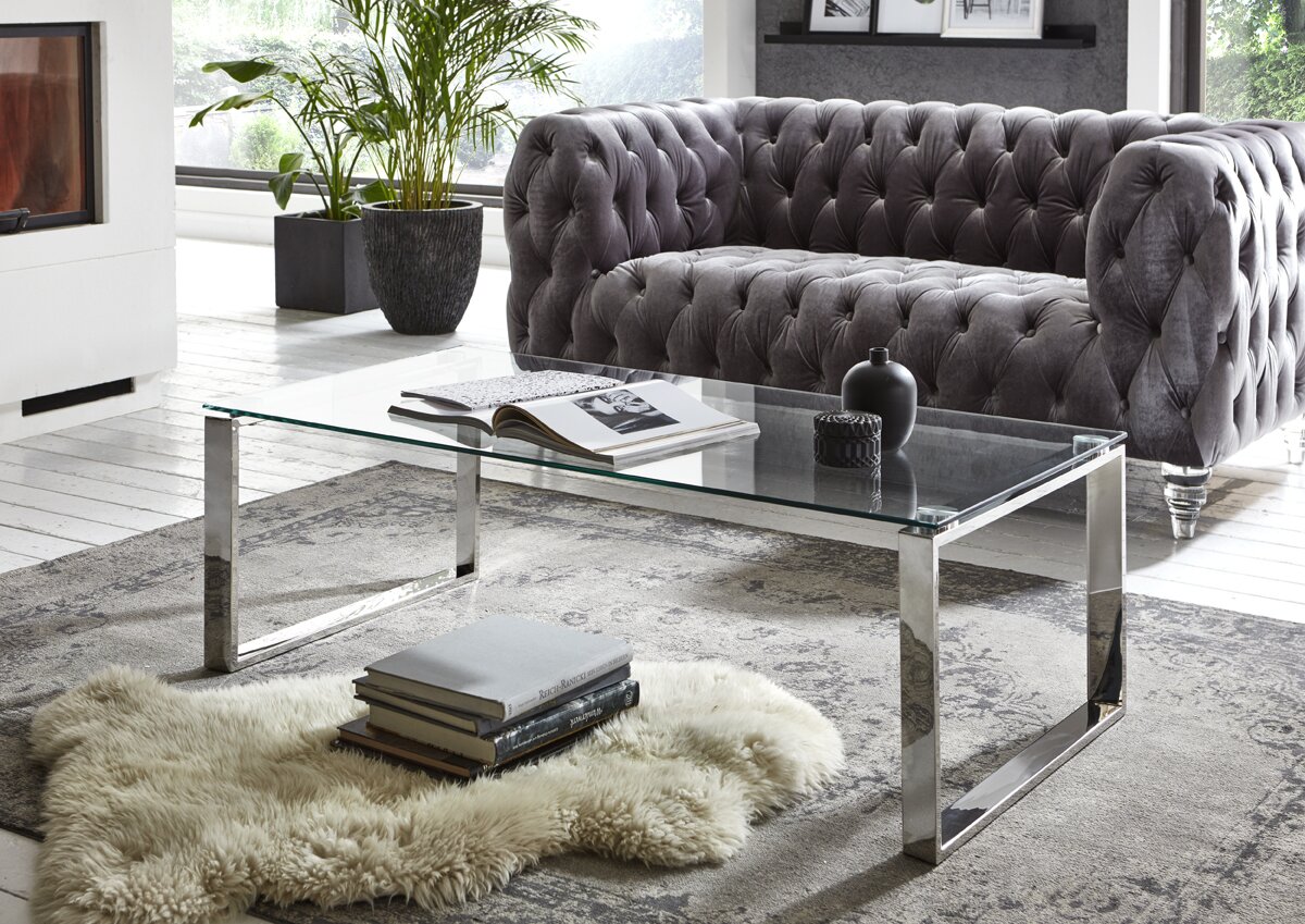 Zurbrüggen Couchtisch Moderner Couchtisch mit transparenter Glasplatte und verchromtem Metallgestell vor grauem Chesterfield-Sofa im Wohnzimmer, Perspektive: leicht schräg von vorne auf Augenhöhe