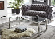 Moderner Couchtisch mit transparenter Glasplatte und verchromtem Metallgestell vor grauem Chesterfield-Sofa im Wohnzimmer, Perspektive: leicht schräg von vorne auf Augenhöhe