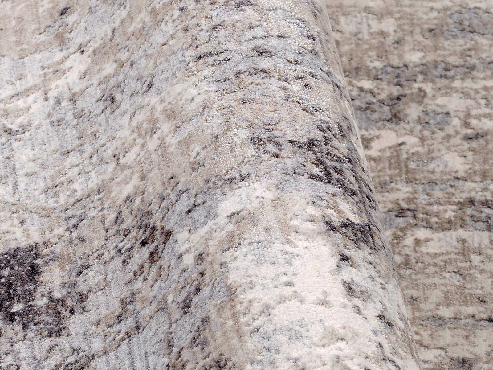 Nahaufnahme eines Designer Teppichs mit abstraktem Muster in Grau- und Beigetönen, leicht gewellt dargestellt.