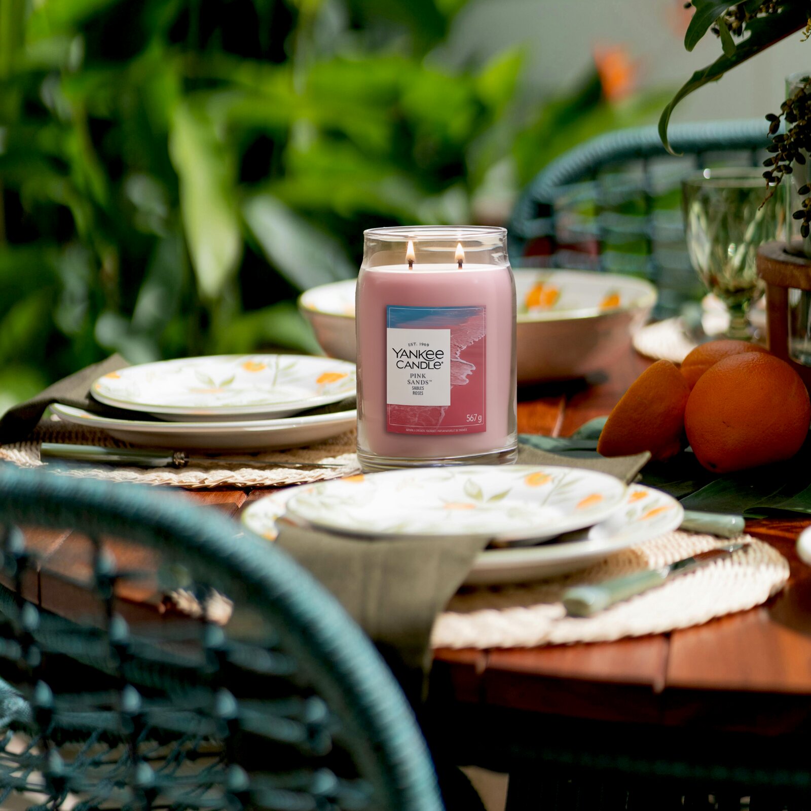 Frontalansicht einer brennenden Yankee Candle Pink Sands Duftkerze im großen Glas mit zwei Dochten auf einem sommerlich gedeckten Gartentisch.