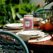 Frontalansicht einer brennenden Yankee Candle Pink Sands Duftkerze im großen Glas mit zwei Dochten auf einem sommerlich gedeckten Gartentisch.