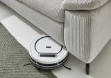 Ein weißer Saugroboter reinigt den Boden unter einem hellgrauen, gerippten Ecksofa. Perspektive von oben.