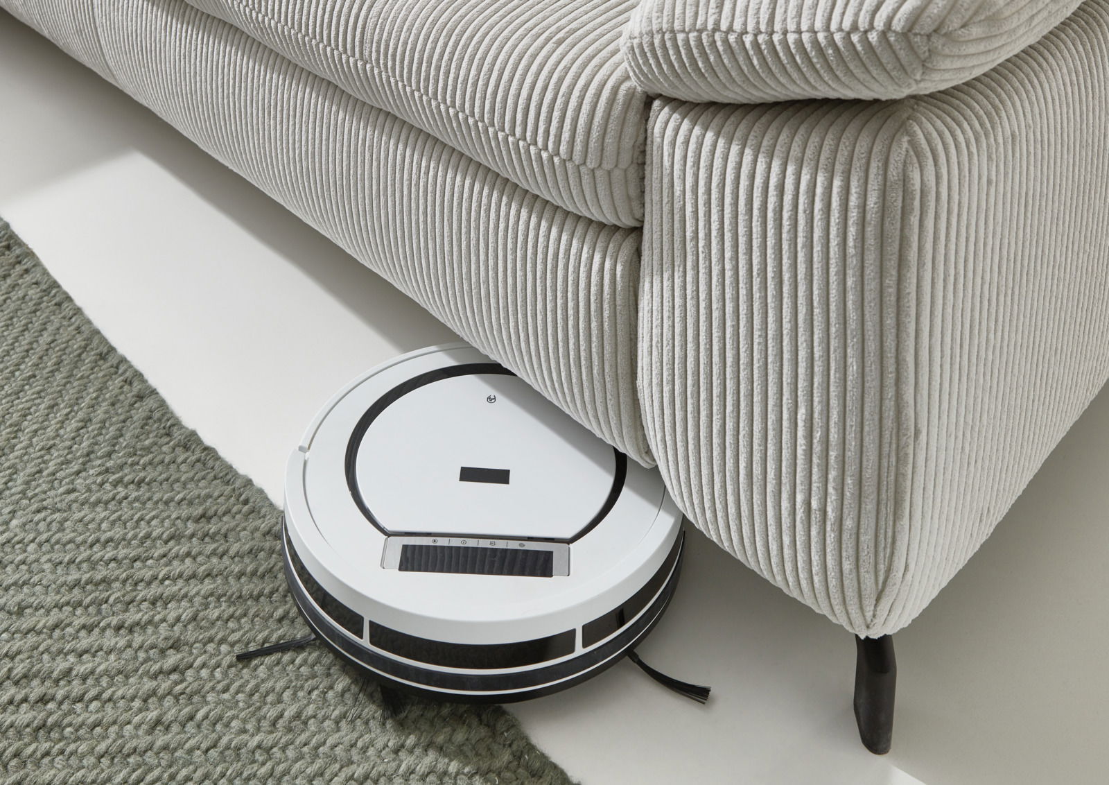 Ein weißer Saugroboter reinigt den Boden unter einem hellgrauen, gerippten Ecksofa. Perspektive von oben.