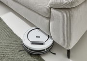 Ein weißer Saugroboter reinigt den Boden unter einem hellgrauen, gerippten Ecksofa. Perspektive von oben.