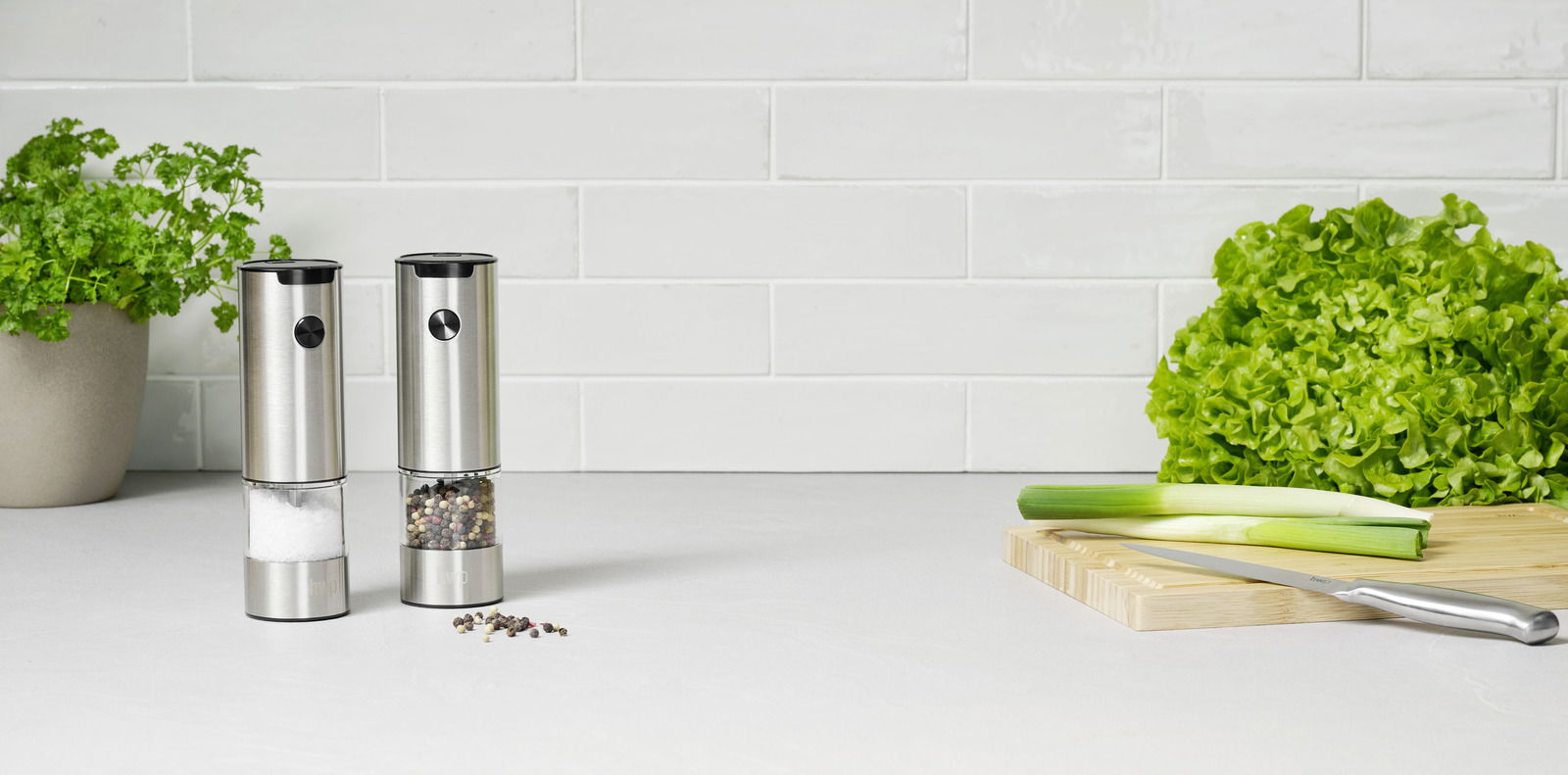 Homeware Salz-/Pfeffermühlen-Set. 2-tlg Salz- und Pfeffermühlen-Set aus Edelstahl, frontal fotografiert auf einer Küchenarbeitsplatte mit frischen Kräutern und Gemüse im Hintergrund.