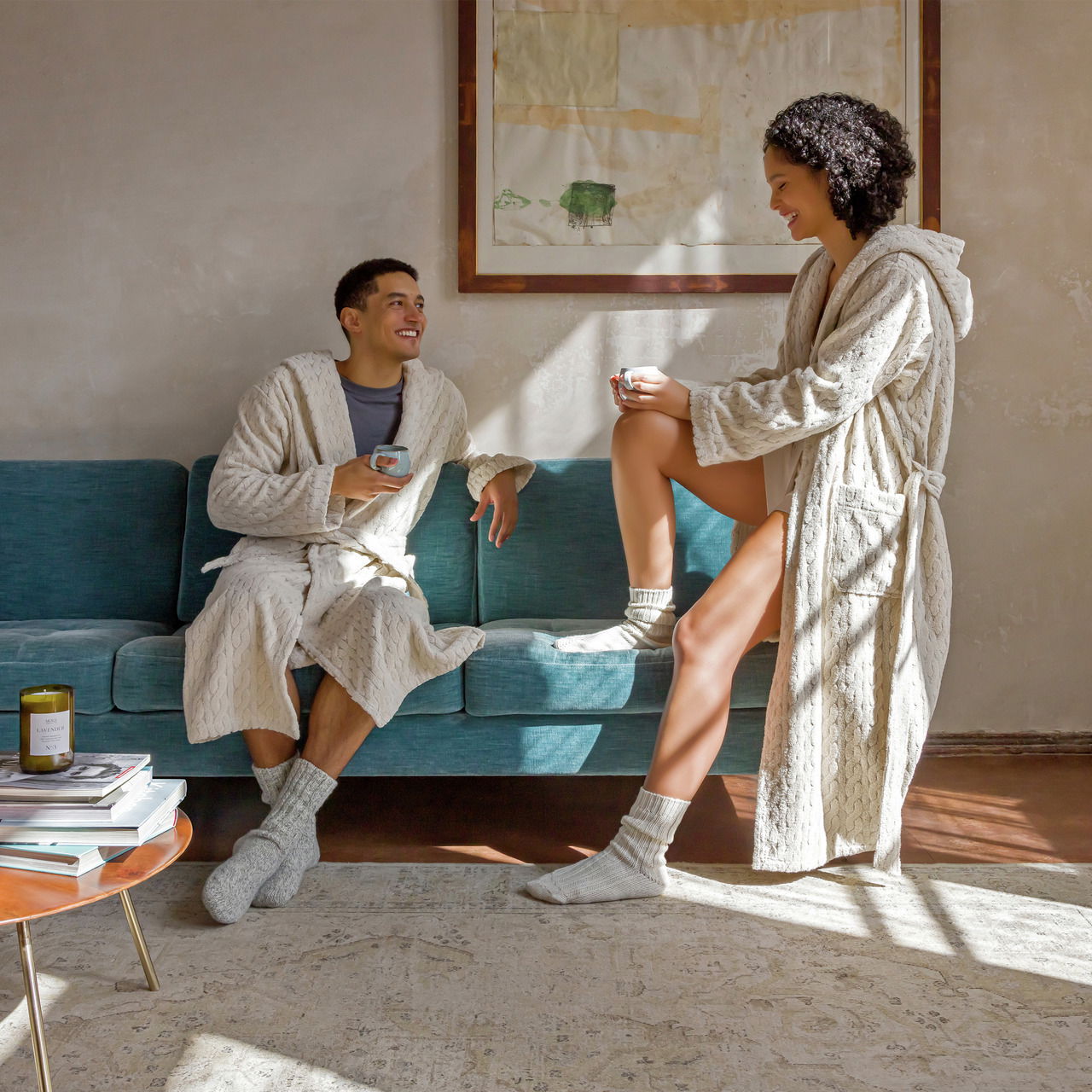 Zwei Personen in beigen Kapuzenmänteln sitzen und stehen auf einem blauen Sofa, genießen eine entspannte Atmosphäre bei Tageslicht.