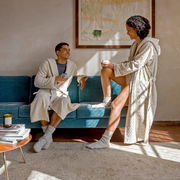 Zwei Personen in beigen Kapuzenmänteln sitzen und stehen auf einem blauen Sofa, genießen eine entspannte Atmosphäre bei Tageslicht.