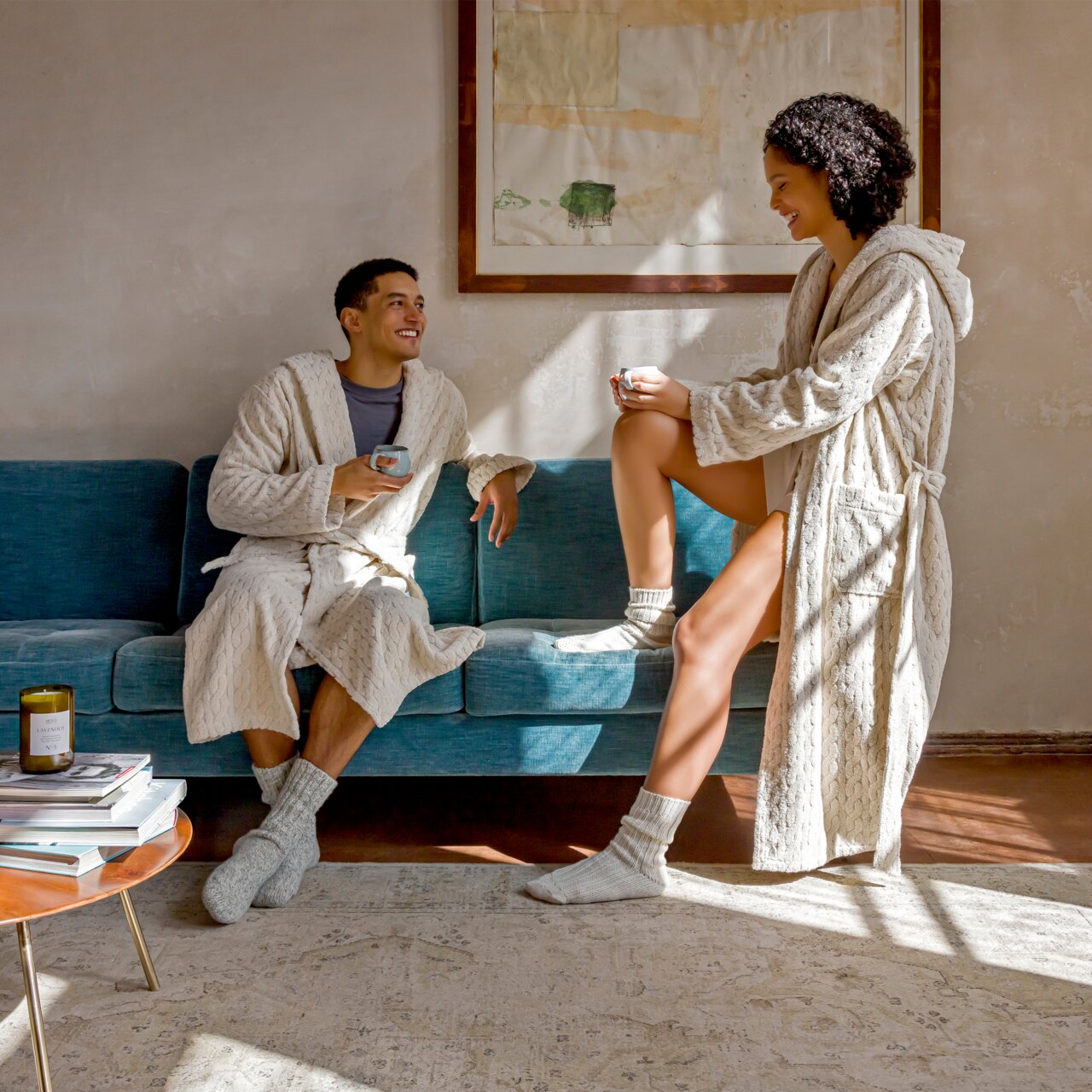 MÖVE Kapuzenmantel, Gr. S COSY KNITS Zwei Personen in beigen Kapuzenmänteln sitzen und stehen auf einem blauen Sofa, genießen eine entspannte Atmosphäre bei Tageslicht.
