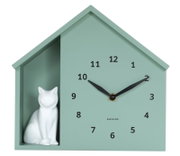 Grüne Wanduhr in Hausform mit schwarzem Zifferblatt und einer weißen Katzenfigur auf der linken Seite, frontal abgebildet.