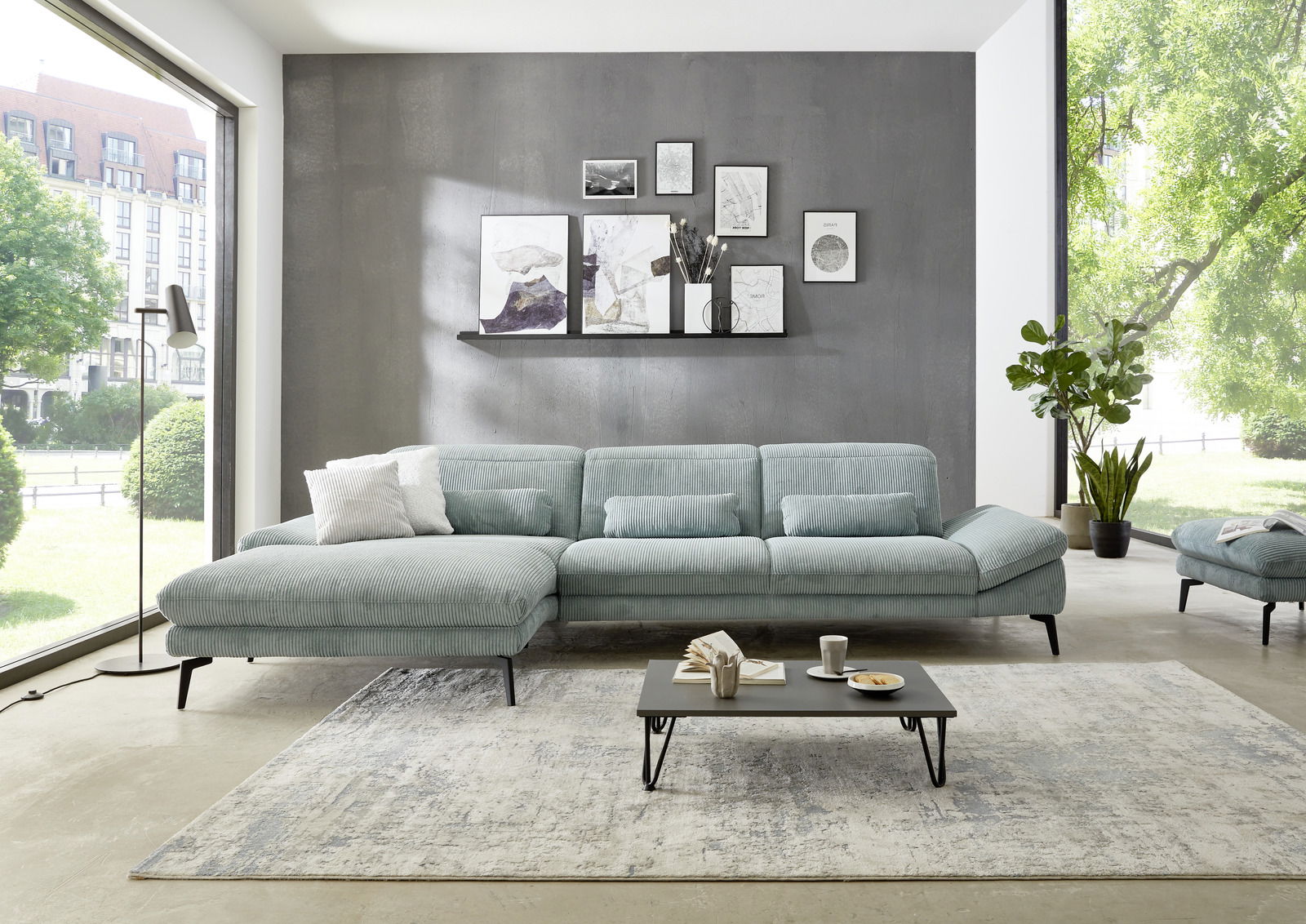 Moderne Longchairkombination in einem hellen Wohnzimmer mit großen Fenstern, grauer Wand und dekorativen Bildern. Perspektive von vorne.