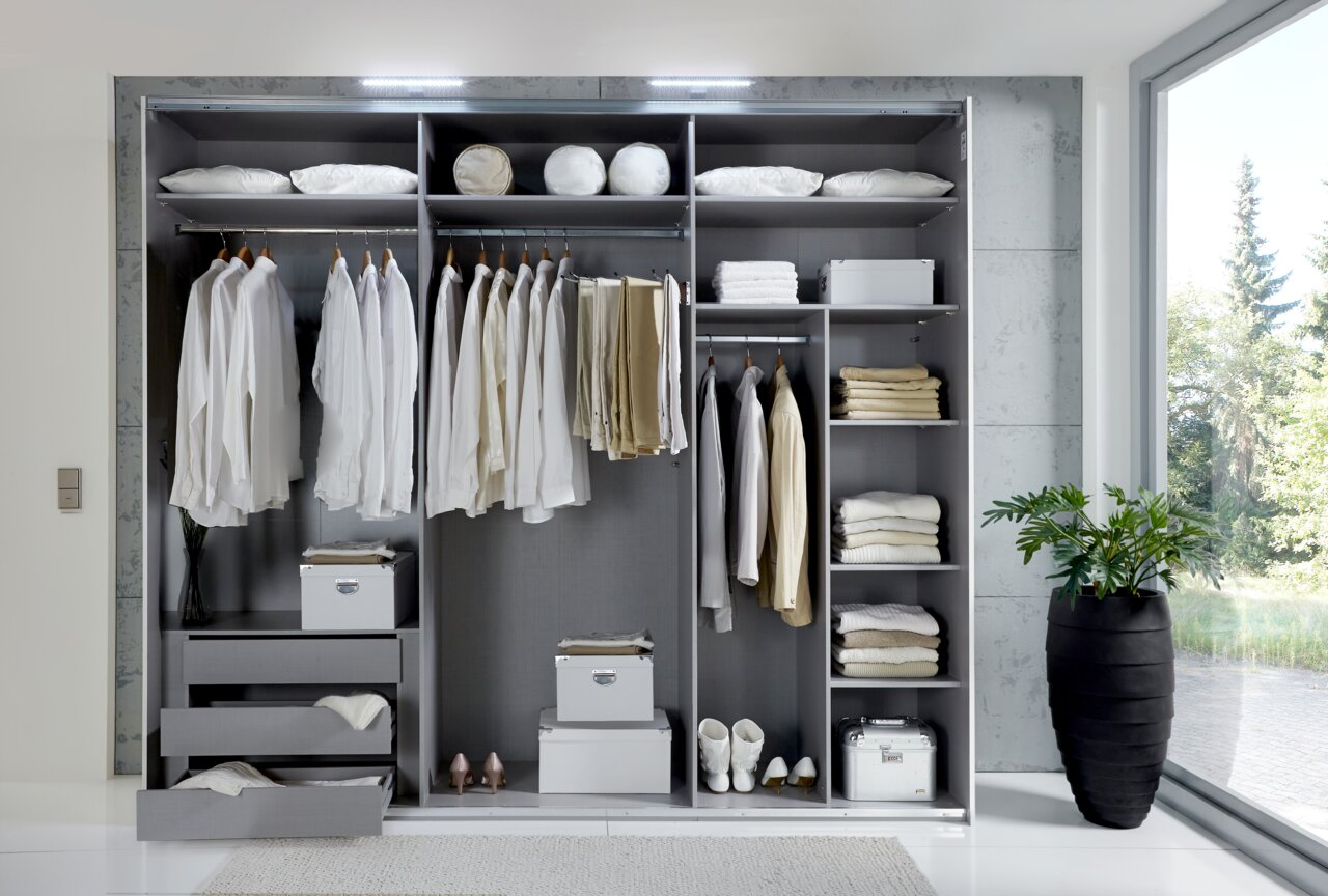 Offener Schwebetürenschrank mit drei Abschnitten, gefüllt mit Kleidung, Kissen und Accessoires, von vorne betrachtet.