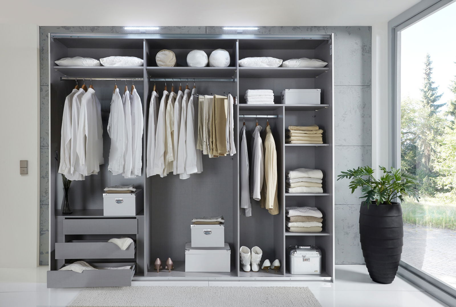 Offener Schwebetürenschrank mit drei Abschnitten, gefüllt mit Kleidung, Kissen und Accessoires, von vorne betrachtet.