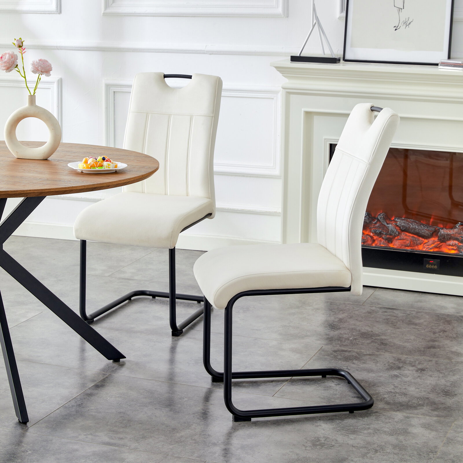 Zwei moderne Esszimmerstühle in Cremeweiß mit gepolsterter Sitz- und Rückenfläche und schwarzem Freischwinger-Metallgestell neben einem runden Holztisch, seitliche Perspektive im hellen Wohnraum mit Kamin im Hintergrund
