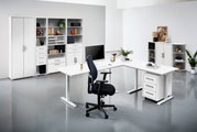 Moderne Büroumgebung mit weißen Schreibtischen, Regalen und einem ergonomischen Bürostuhl, aus der Perspektive von vorne betrachtet.