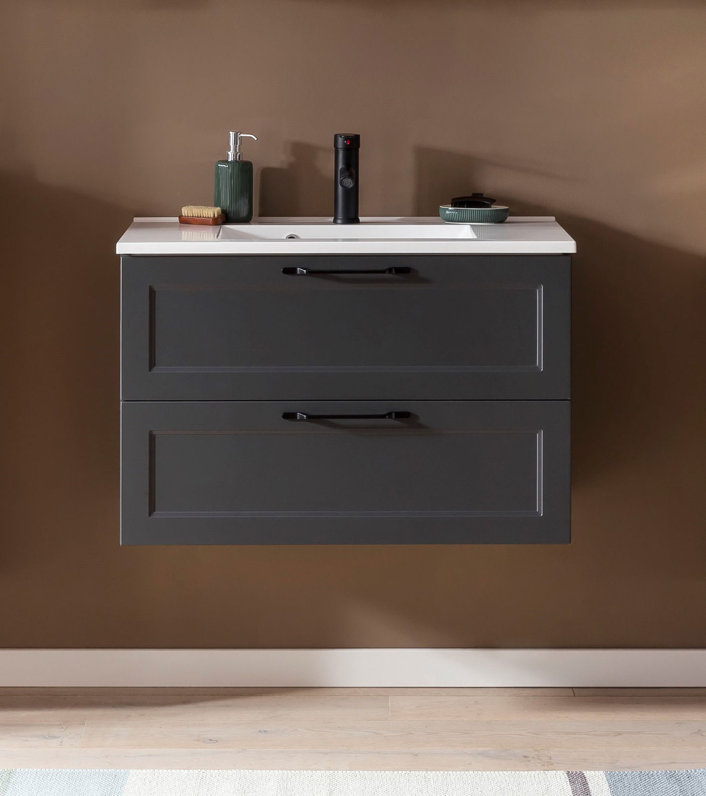 Frontale Ansicht eines modernen Waschtisch-Sets mit zwei Schubladen in dunklem Grau, weißer Waschbeckenoberfläche und stilvollen schwarzen Griffen, dekoriert mit Seifenspender und Accessoires.