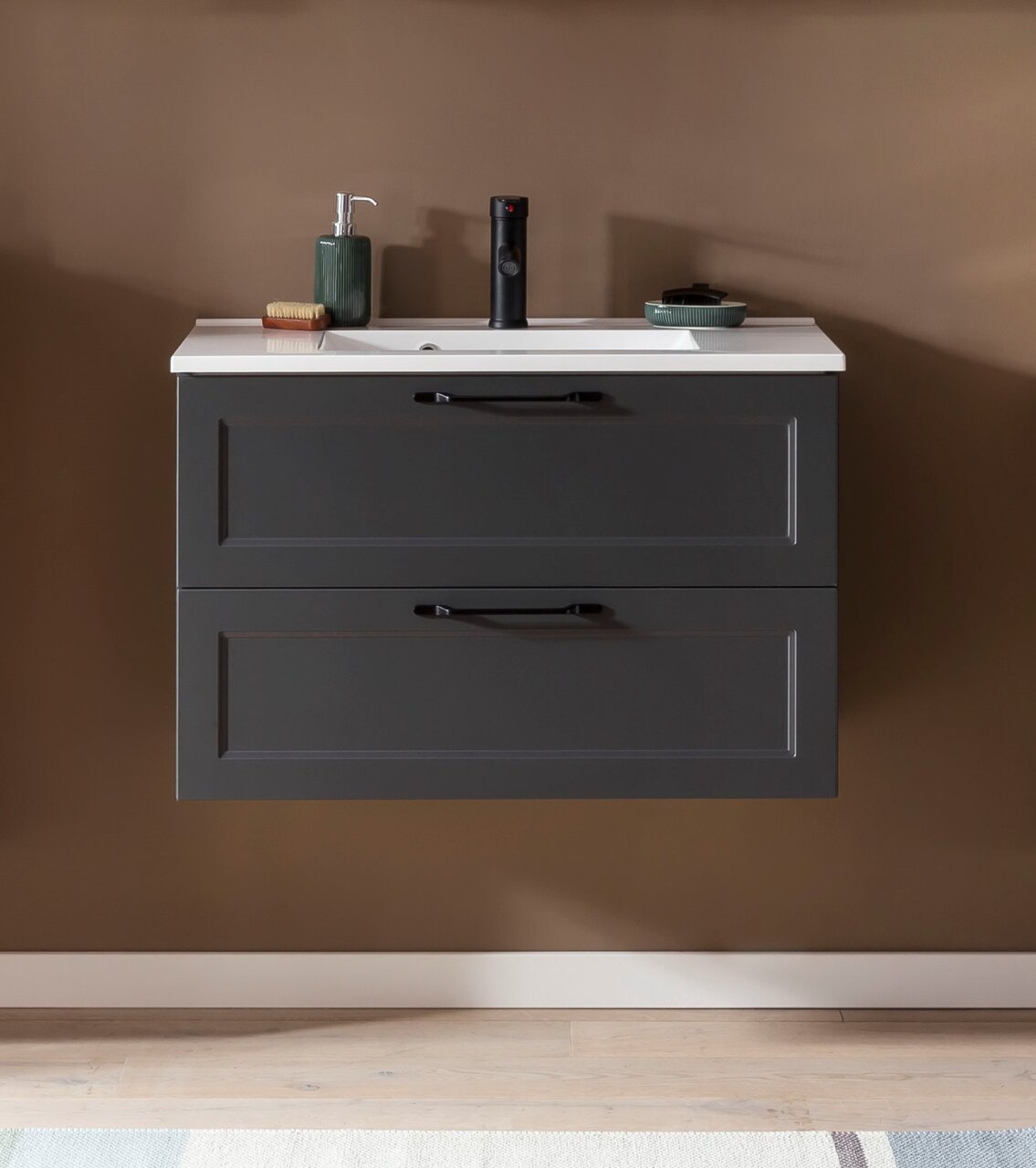 Vooraanzicht van een moderne wastafelcombinatie met twee lades in donkergrijs, een wit wastafelblad en stijlvolle zwarte handgrepen, aangevuld met een zeepdispenser en accessoires.