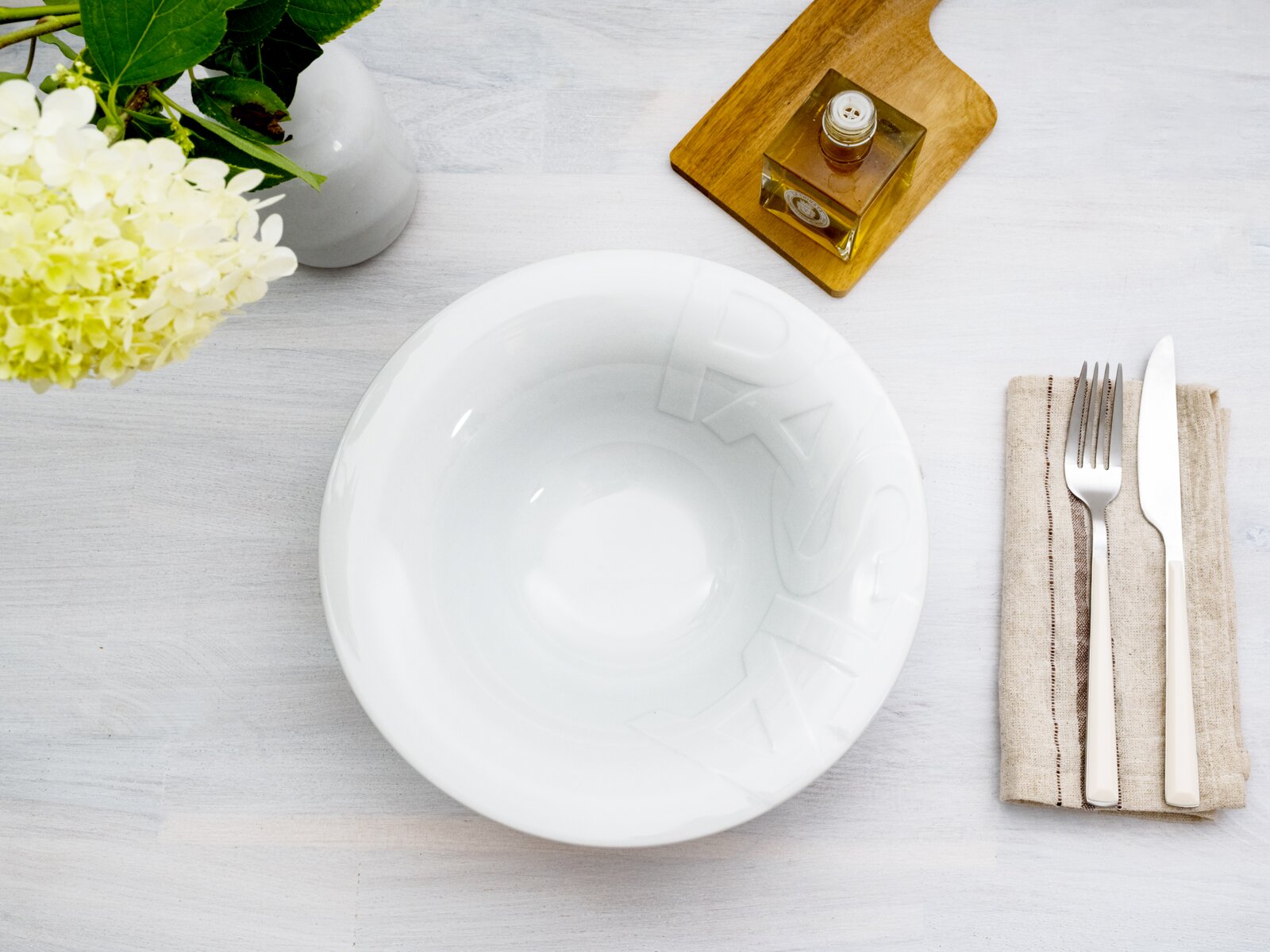 Weiße, glänzende Schale mit dezenter Reliefschrift, Draufsicht auf weißer Holzoberfläche, daneben Besteck auf Stoffserviette sowie Vase mit Blumen und kleines Glasgefäß auf Holzbrett
