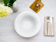 Weiße, glänzende Schale mit dezenter Reliefschrift, Draufsicht auf weißer Holzoberfläche, daneben Besteck auf Stoffserviette sowie Vase mit Blumen und kleines Glasgefäß auf Holzbrett