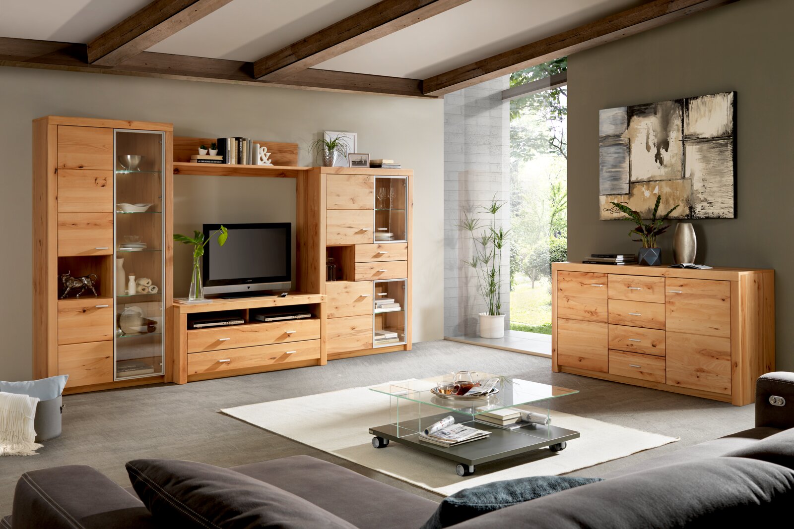 Cantus Dressoir INDIRA Schuine frontaanzicht van een moderne woonkamer met lichte houten wandmeubelset met vitrinekasten en tv-meubel, aangevuld met een bijpassende dressoir en een glazen salontafel op wielen.