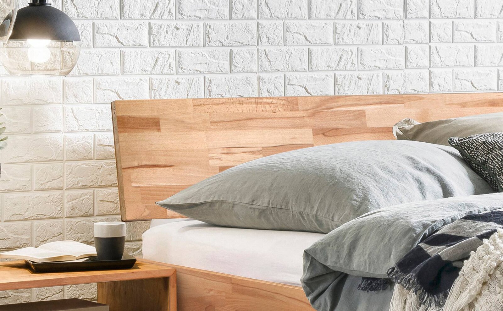 Houten bedframe met hoofdeinde van licht hout, gezien vanaf de zijkant, gedecoreerd met grijs beddengoed en een nachtkastje met lamp.