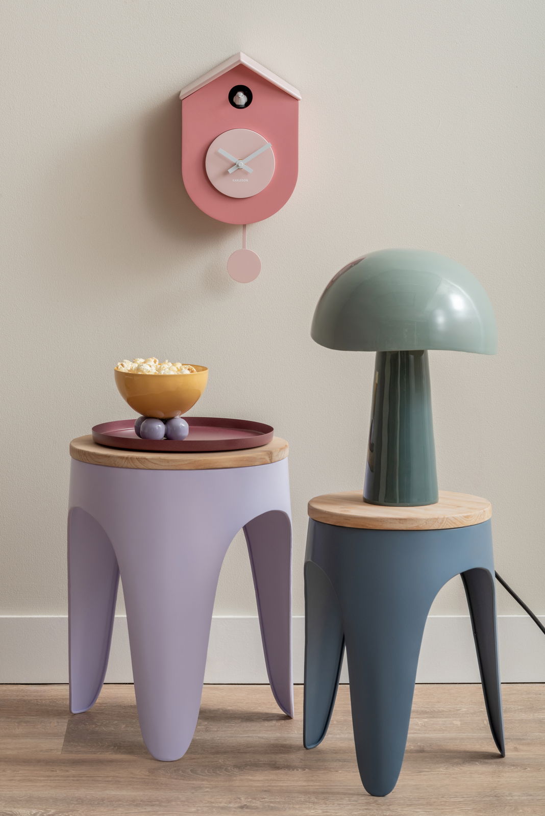 Rosa Wanduhr in Vogelhausform mit Pendel, frontal an der Wand montiert, neben modernen Beistelltischen und einer Lampe.