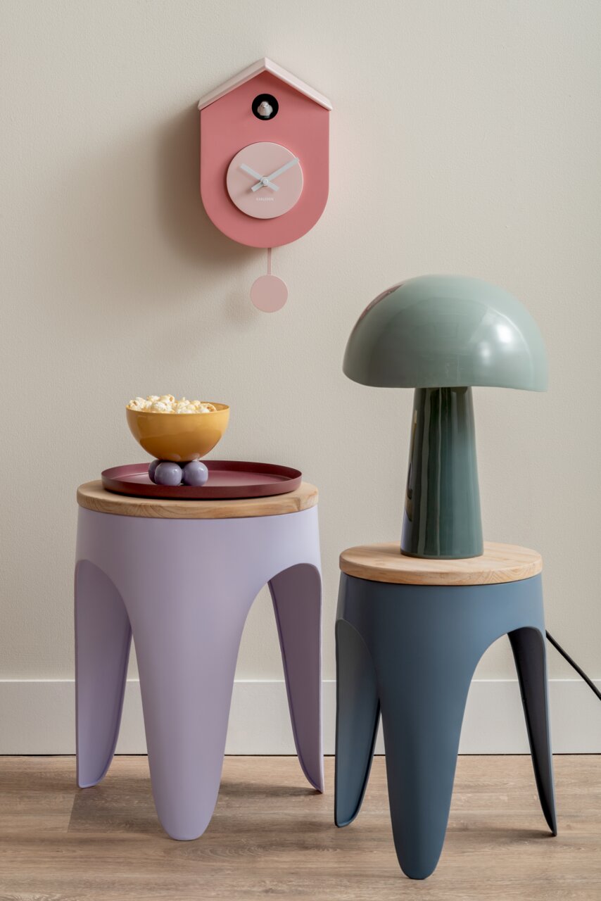 KARLSSON Wandklok Grato CUCKOO Roze wandklok in de vorm van een vogelhuisje met slinger, aan de voorkant aan de muur bevestigd, naast moderne bijzettafeltjes en een lamp.