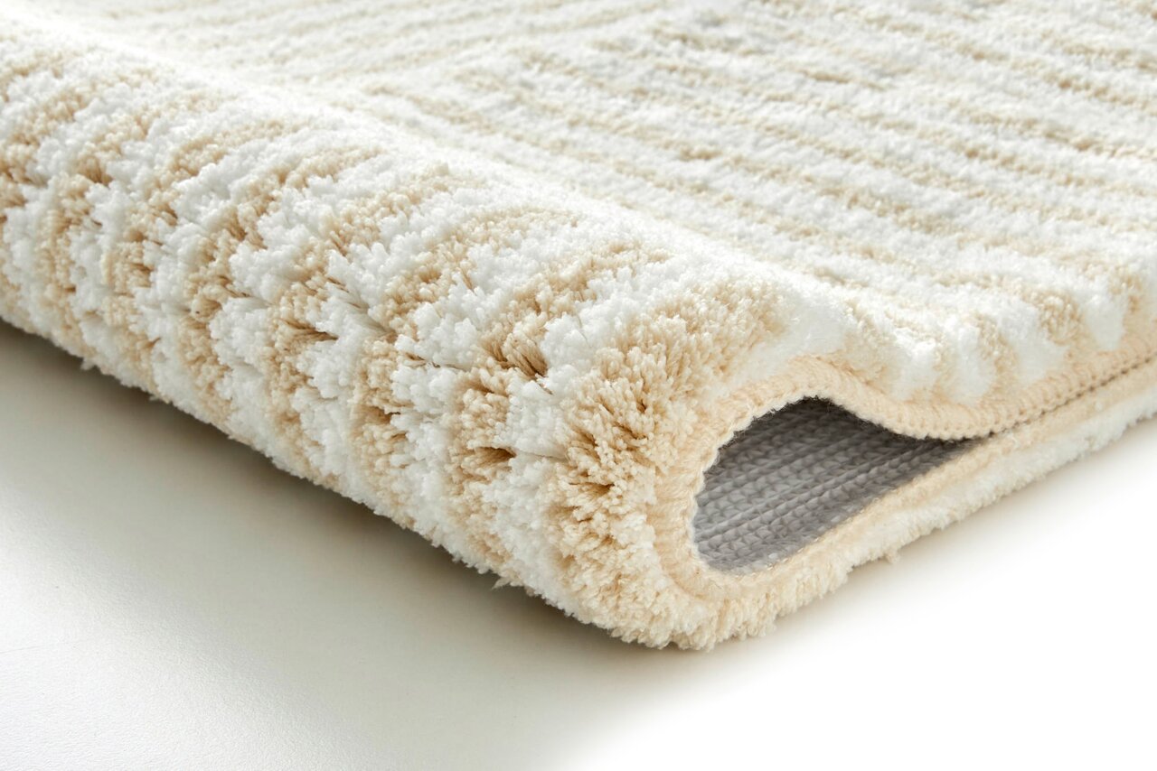 Nahaufnahme eines beigen und weißen Badteppichs mit flauschiger Textur, seitliche Perspektive