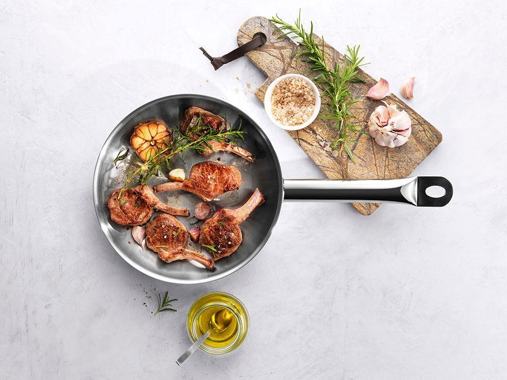 Bratpfanne mit gebratenen Koteletts, Knoblauch und Kräutern, daneben Gewürze und Olivenöl.