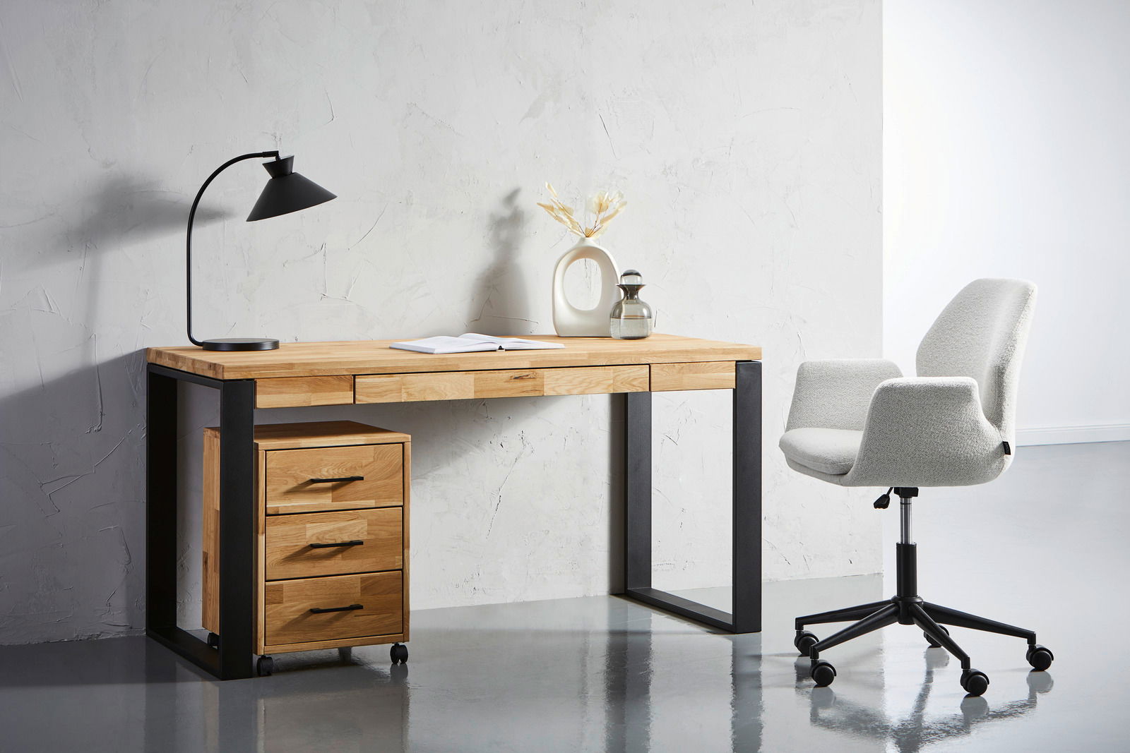 Moderner Schreibtisch aus Holz mit schwarzem Metallgestell, links mit Schubladencontainer auf Rollen, daneben ein grauer Bürostuhl. Perspektive von vorne.