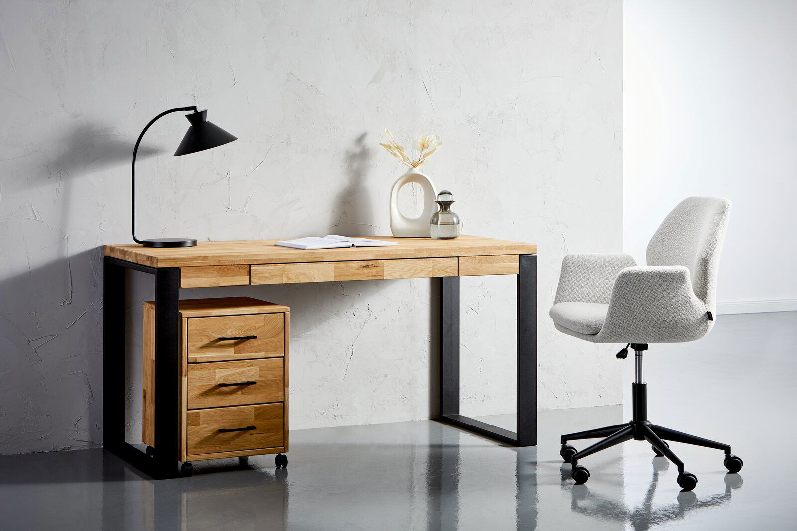 Moderner Schreibtisch aus Holz mit schwarzem Metallgestell, links mit Schubladencontainer auf Rollen, daneben ein grauer Bürostuhl. Perspektive von vorne.