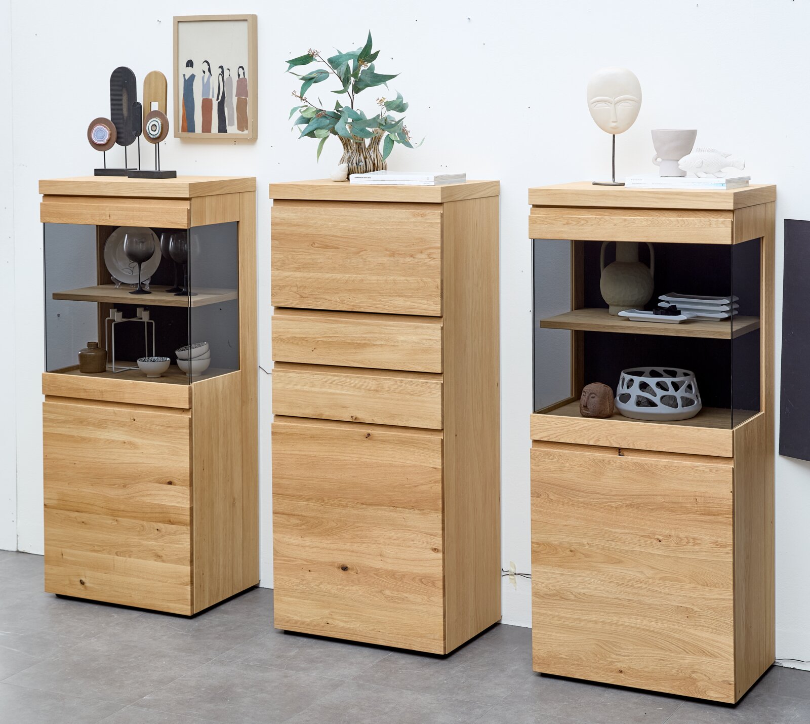 Drei moderne Hochschränke aus naturbelassenem Holz, zwei davon mit verglasten Vitrinenfächern und dekorativen Wohnaccessoires, in frontaler Perspektive mit leicht sichtbaren Seitenflächen.