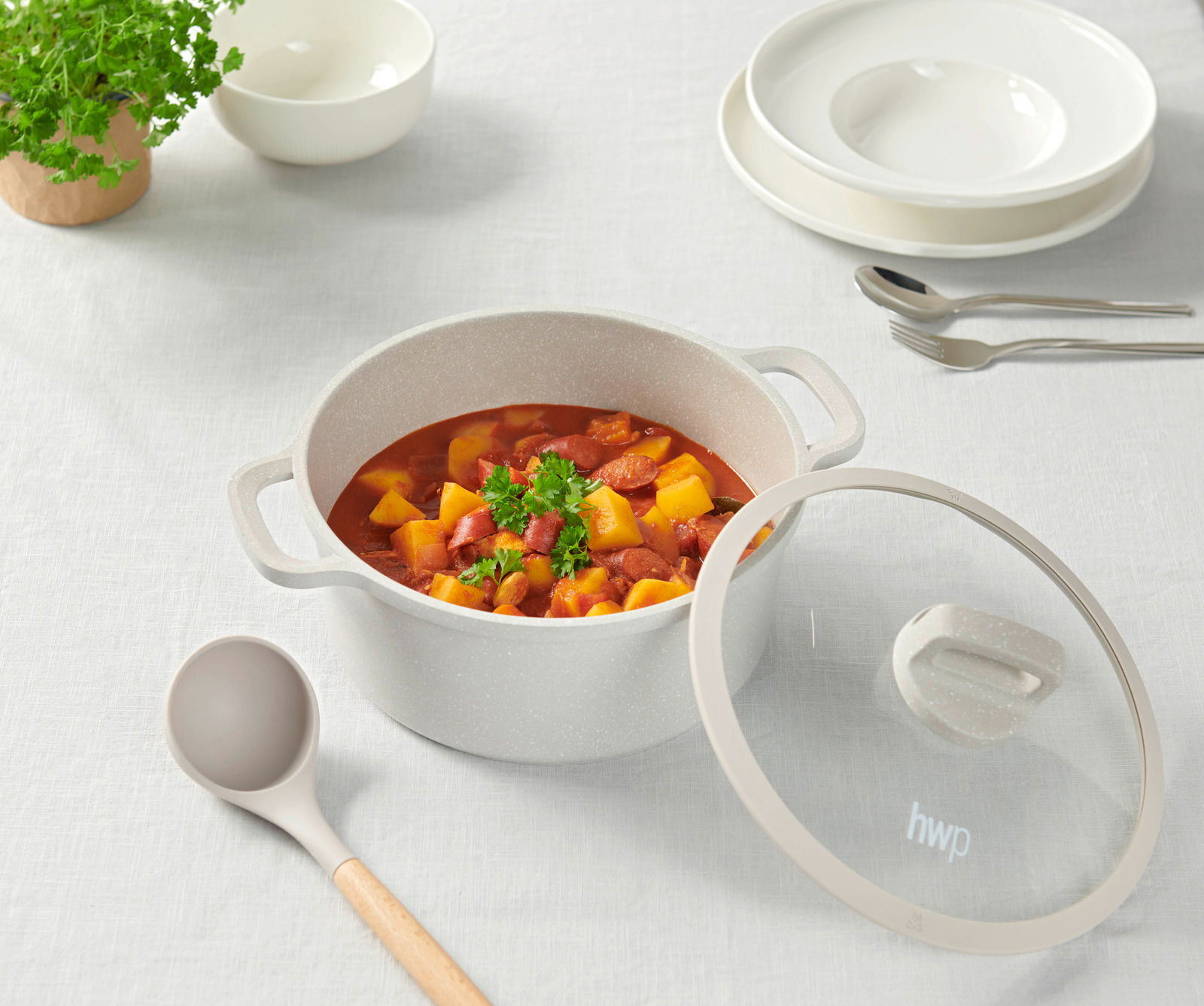 Ein Kochtopf mit Deckel, gefüllt mit einem Eintopf aus Gemüse und Wurst, auf einem Tisch mit weißem Tischtuch. Perspektive von oben.