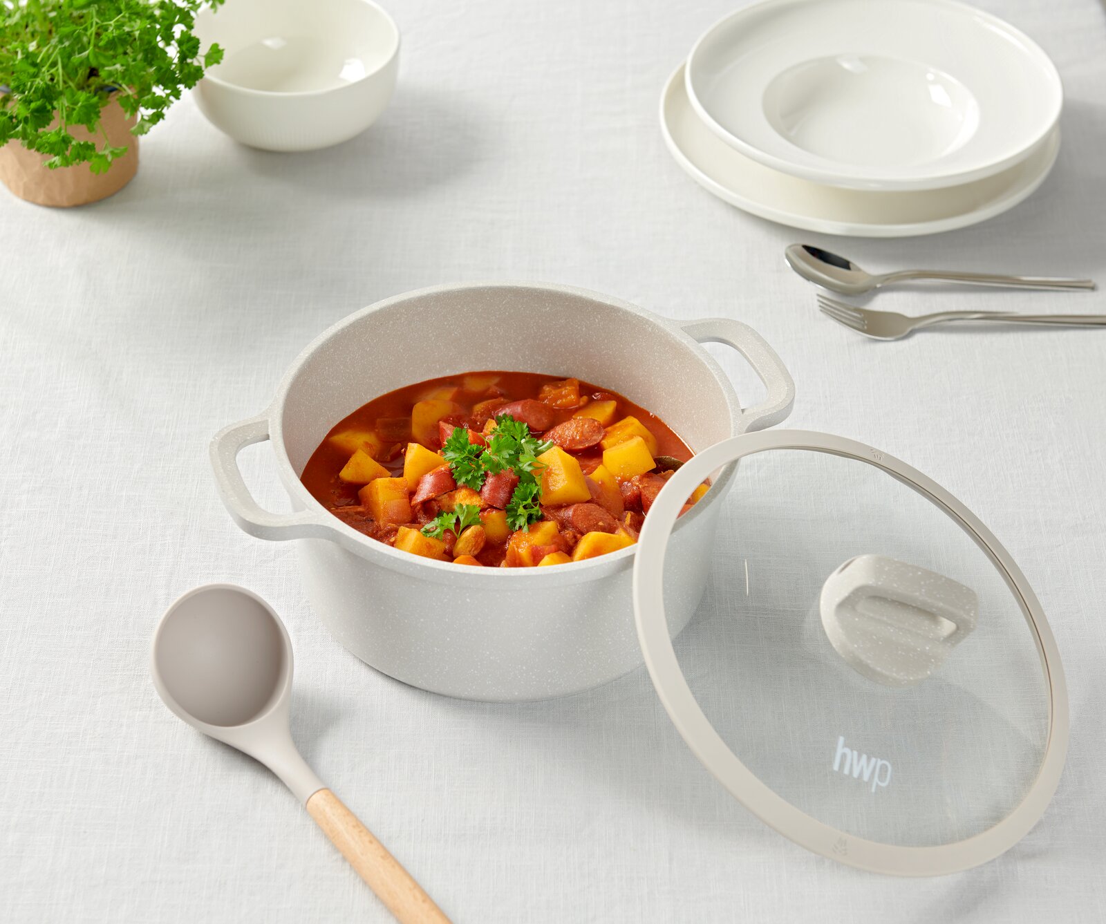 Ein Kochtopf mit Deckel, gefüllt mit einem Eintopf aus Gemüse und Wurst, auf einem Tisch mit weißem Tischtuch. Perspektive von oben.