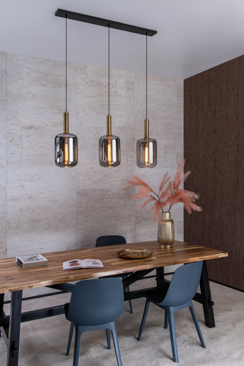 Drei moderne Pendelleuchten JOANET mit rauchgrauen Glaskörpern und goldenen Fassungen, hängend über einem Esstisch in einem stilvoll eingerichteten Raum. Seitliche Perspektive.