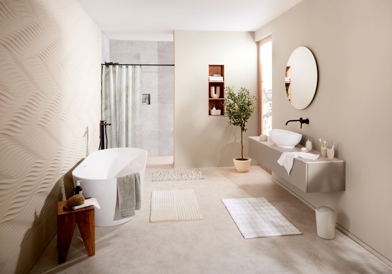 Modernes Badezimmer mit freistehender Badewanne, mehreren Badteppichen in neutralen Farben und einem großen Spiegel, seitliche Perspektive.