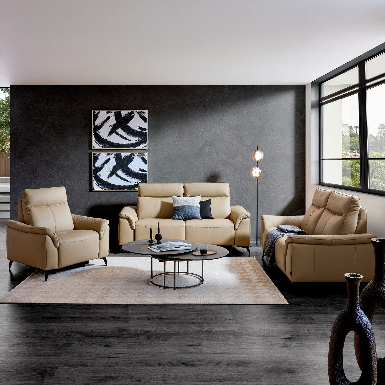 Wohnzimmeransicht mit beigem Ledersofa-Set bestehend aus einem 3-Sitzer, einem 2-Sitzer und einem Sessel, vor einer grauen Wand mit moderner Kunst, auf einem hellen Teppich mit Couchtisch und Stehlampe.