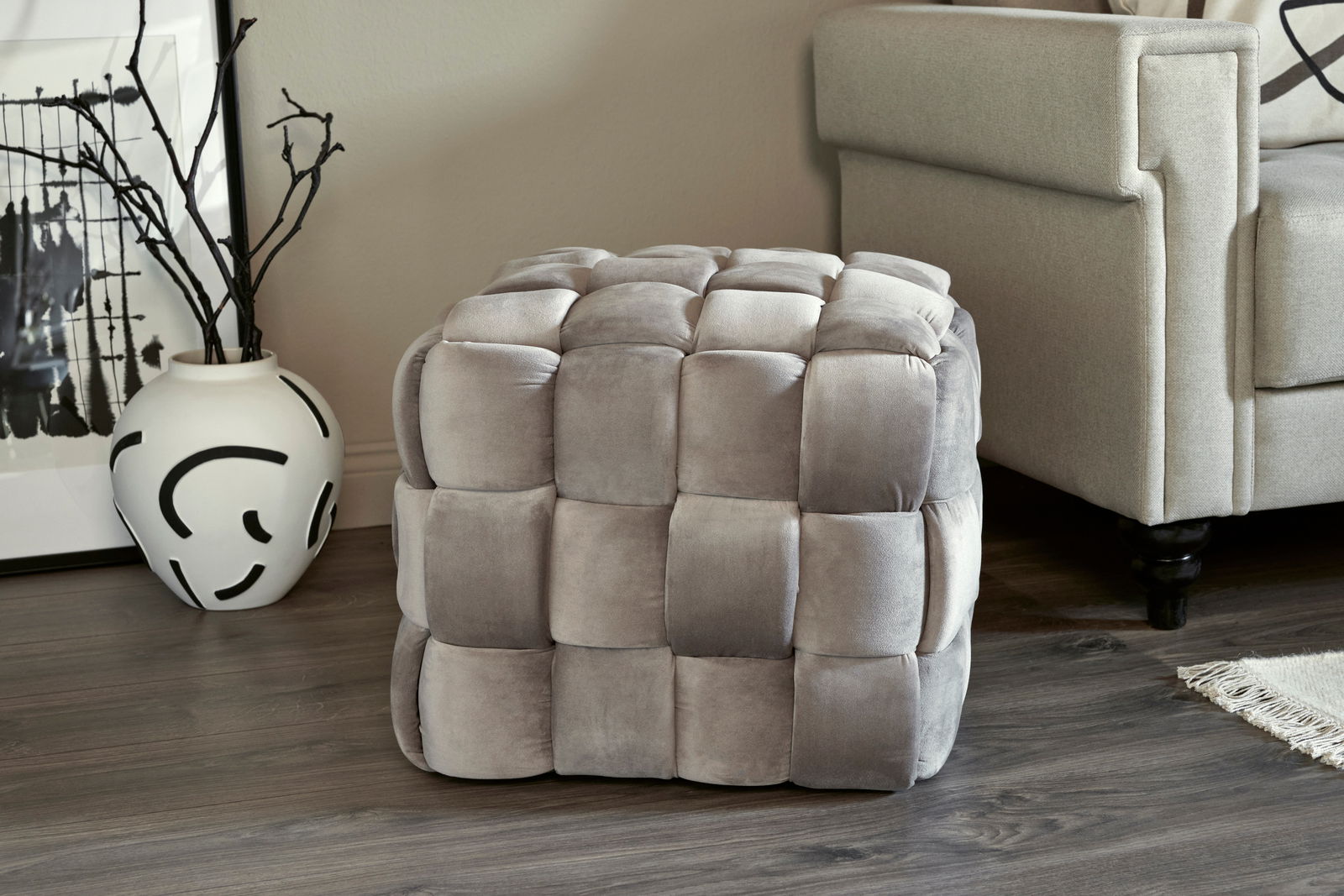 Grauer, gepolsterter Hocker in Würfelform mit quadratischem Muster, seitliche Perspektive.