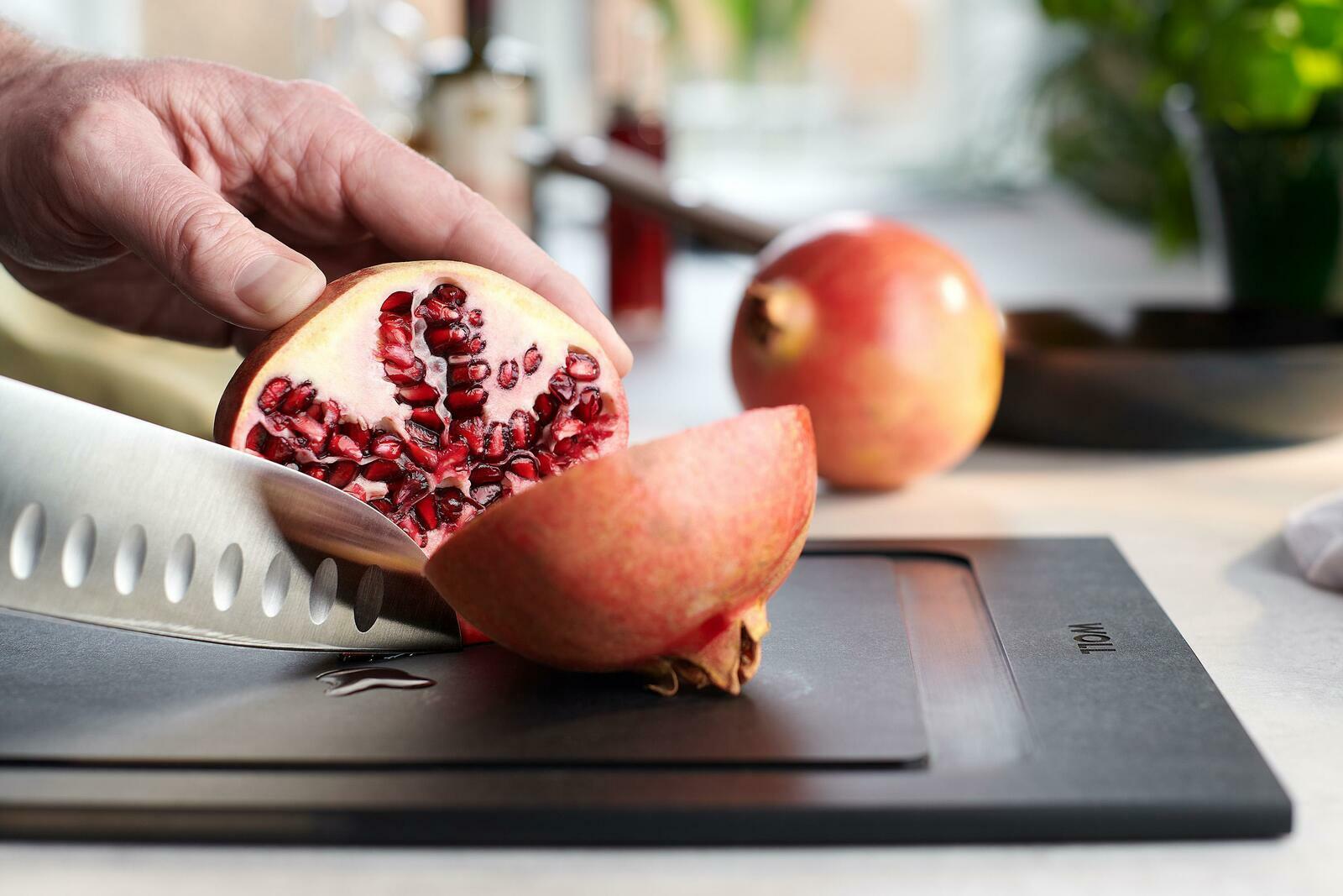 Nahaufnahme eines Schneidebretts mit einem aufgeschnittenen Granatapfel und einem Messer, das von einer Hand gehalten wird. Perspektive von der Seite.