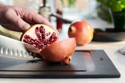 Nahaufnahme eines Schneidebretts mit einem aufgeschnittenen Granatapfel und einem Messer, das von einer Hand gehalten wird. Perspektive von der Seite.