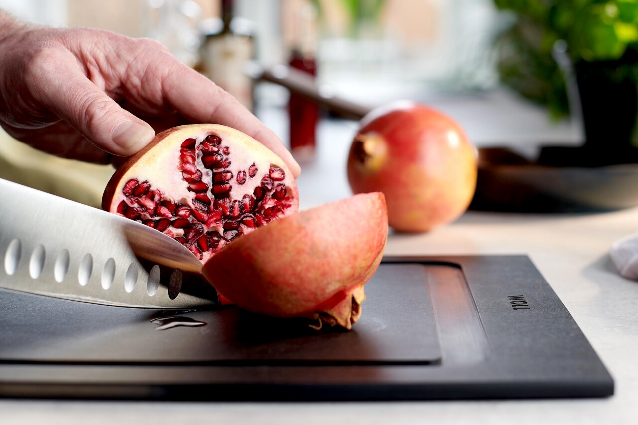 Nahaufnahme eines Schneidebretts mit einem aufgeschnittenen Granatapfel und einem Messer, das von einer Hand gehalten wird. Perspektive von der Seite.