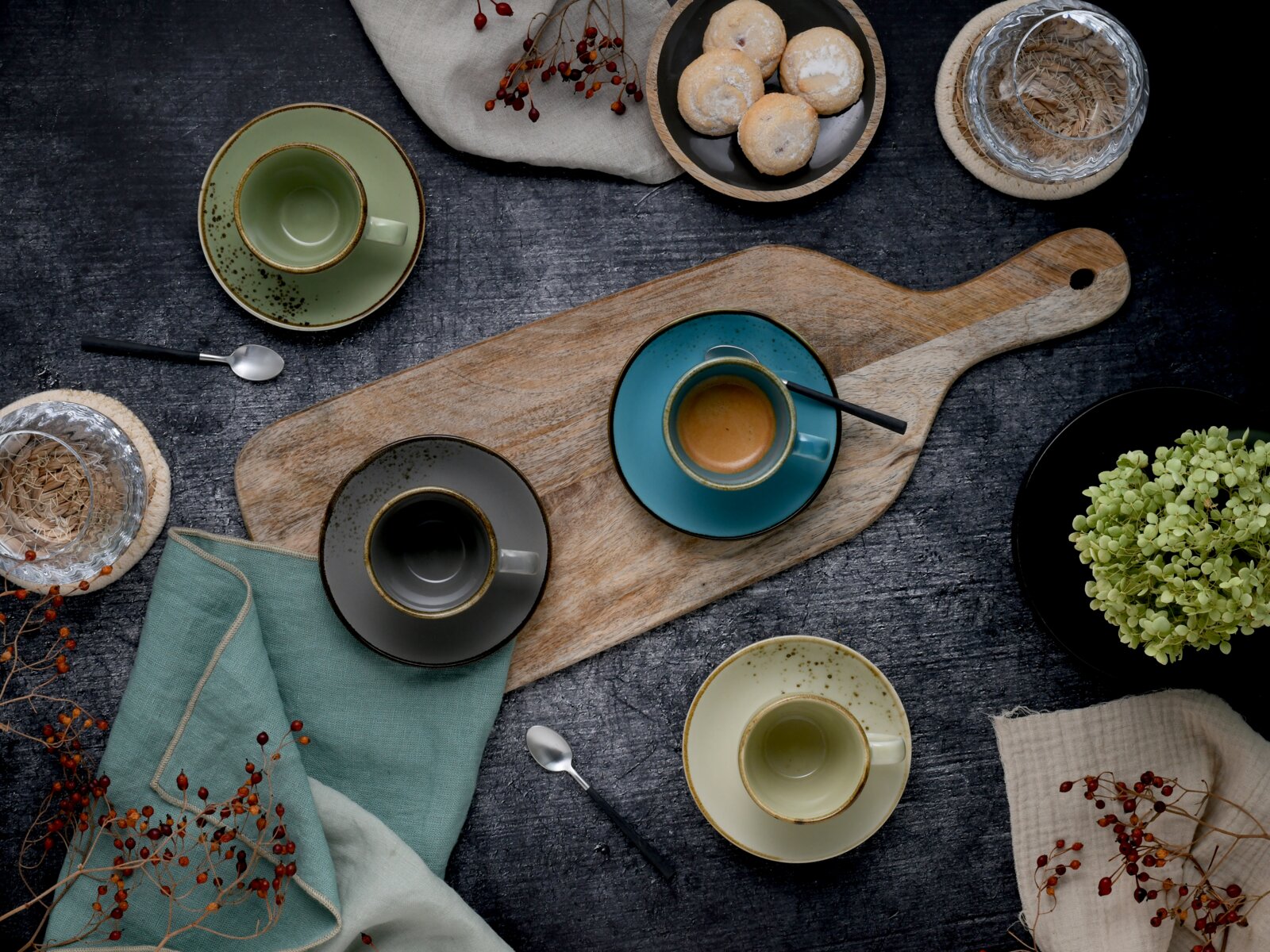 Draufsicht (Flatlay) auf rustikale Kaffeetafel mit mehreren farbigen Espresso- und Kaffeetassen auf Holzbrett, Löffeln, Gebäck, Gläsern und Deko auf dunklem Tisch